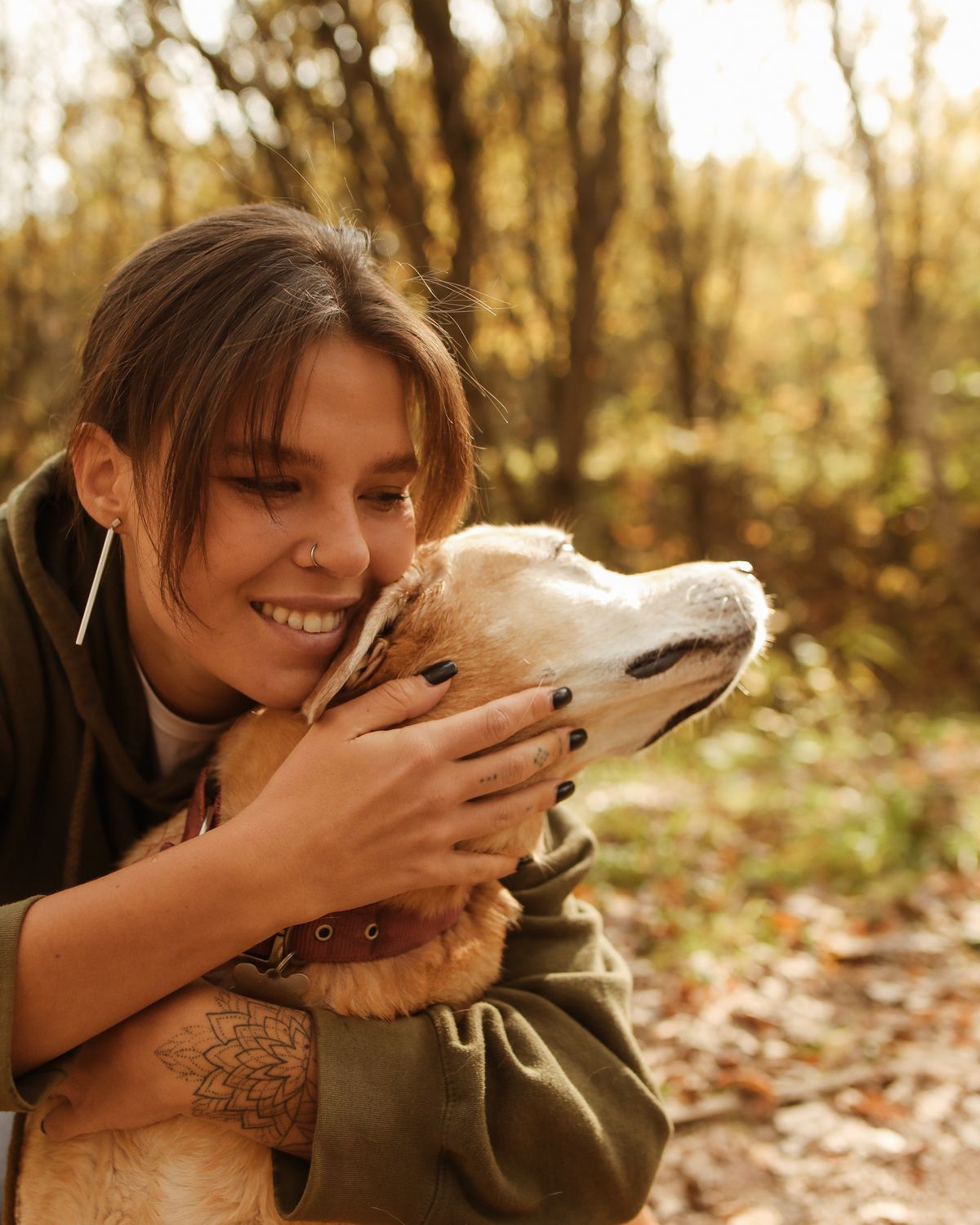 Une jeune femme souriante serre son chien dans ses bras, dans une forêt baignée de soleil d'automne